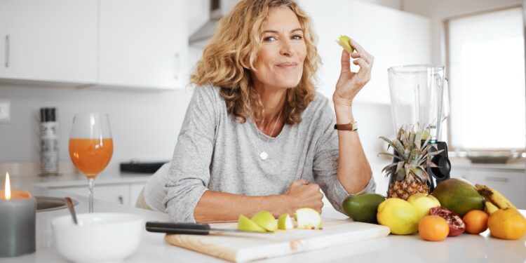 Smiling woman leans at a kitchen counter with cut fruit, a blender, citrus, pineapple, bananas, and juice.